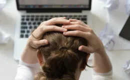 Mulher segurando a cabeça com as mãos, simbolizando estresse e sobrecarga mental diante de um computador, cercada por papéis amassados.