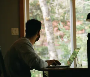 Um homem sentado à mesa com um notebook/laptop olhando para o lado de fora através da janela